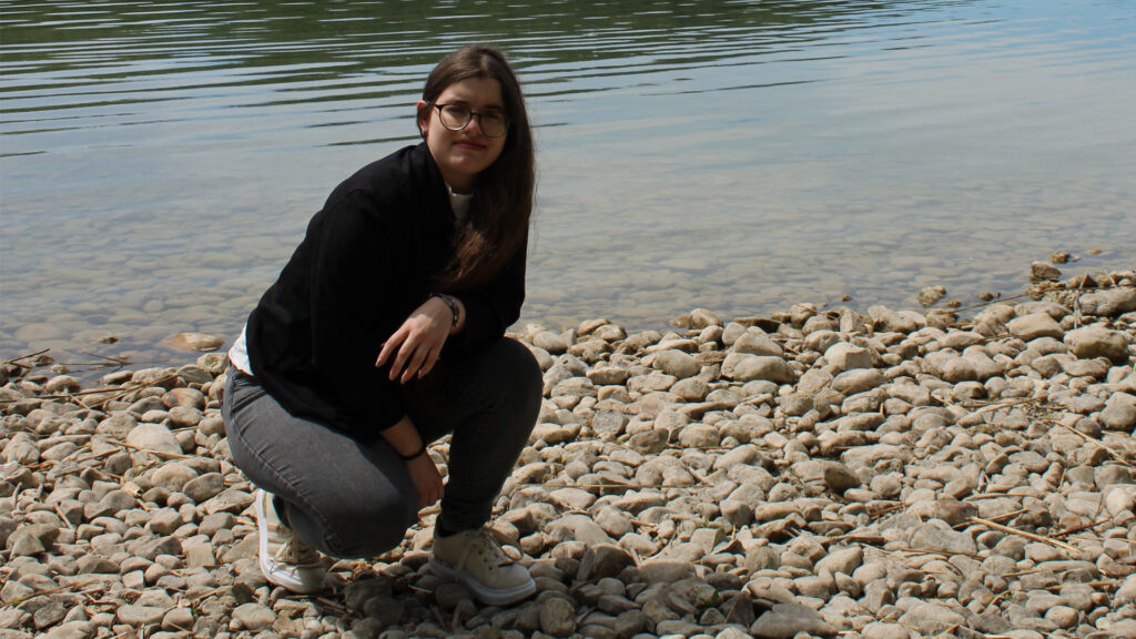Ganzkörperporträt von mir in hockender Position. Im Hintergrund ist ein See und ein Steinufer zu sehen.
