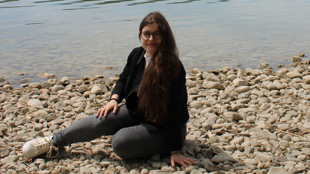Ganzkörperporträt von mir in sitzender Position. Im Hintergrund ist ein See und ein Steinufer zu sehen.
