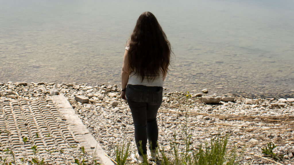Ganzkörperporträt von mir in stehen mit Blick zum Wasser. Im Hintergrund ist ein See und ein Steinufer zu sehen.