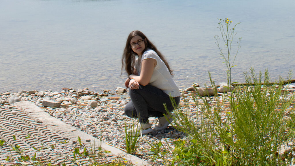 Ganzkörperporträt von mir in hockender Position. Im Hintergrund ist ein See und ein Steinufer zu sehen.